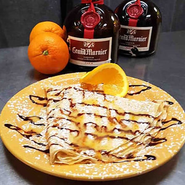 Crepe Suzette on a yellow plate, garnished with an orange slice, powdered sugar, and chocolate drizzle.